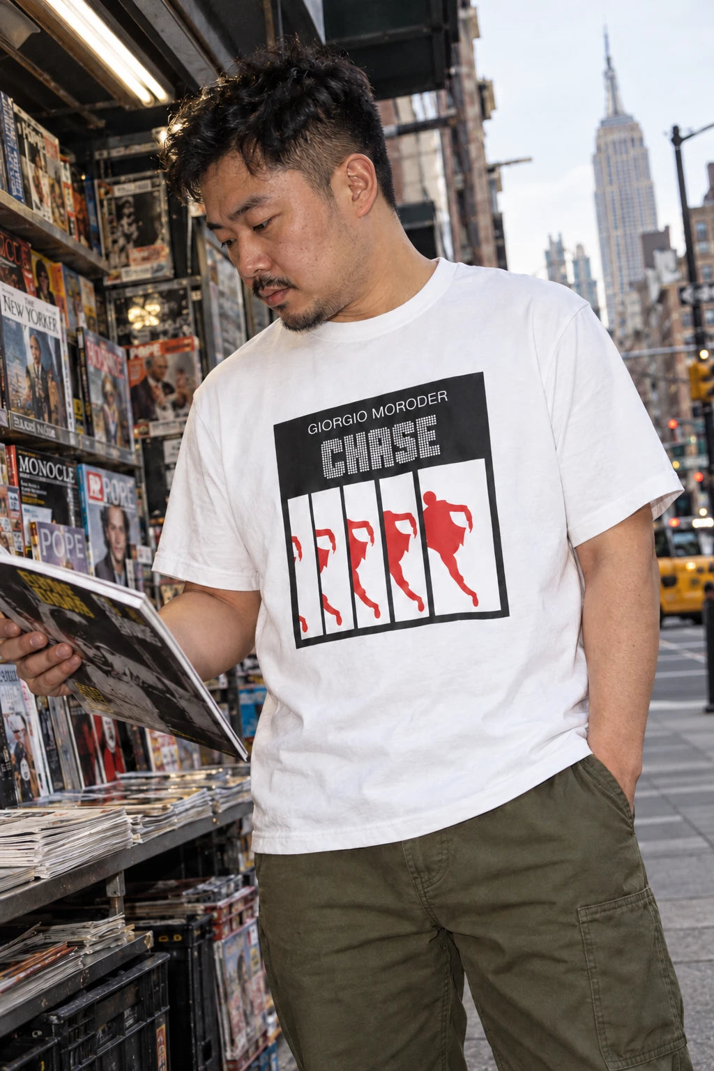 Man wearing a white Giorgio Moroder Chase T-shirt while browsing a magazine at a city newsstand.