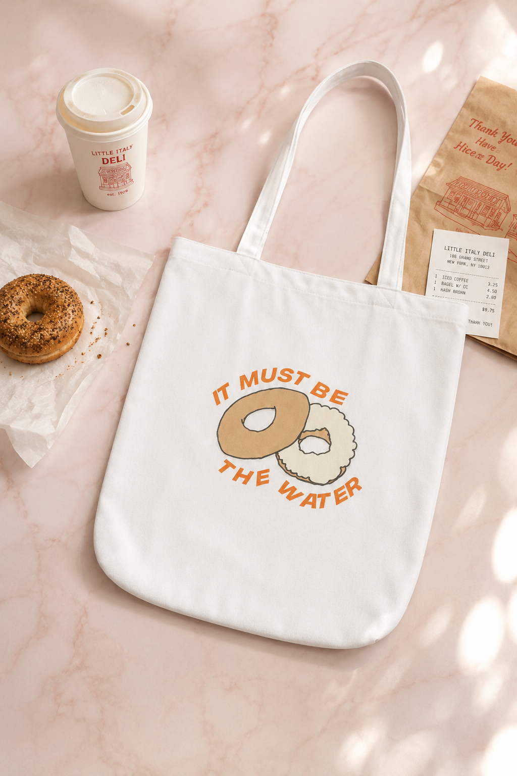Hand-drawn It Must Be The Water bagel tote bag styled in a soft New York deli-inspired editorial flat lay.