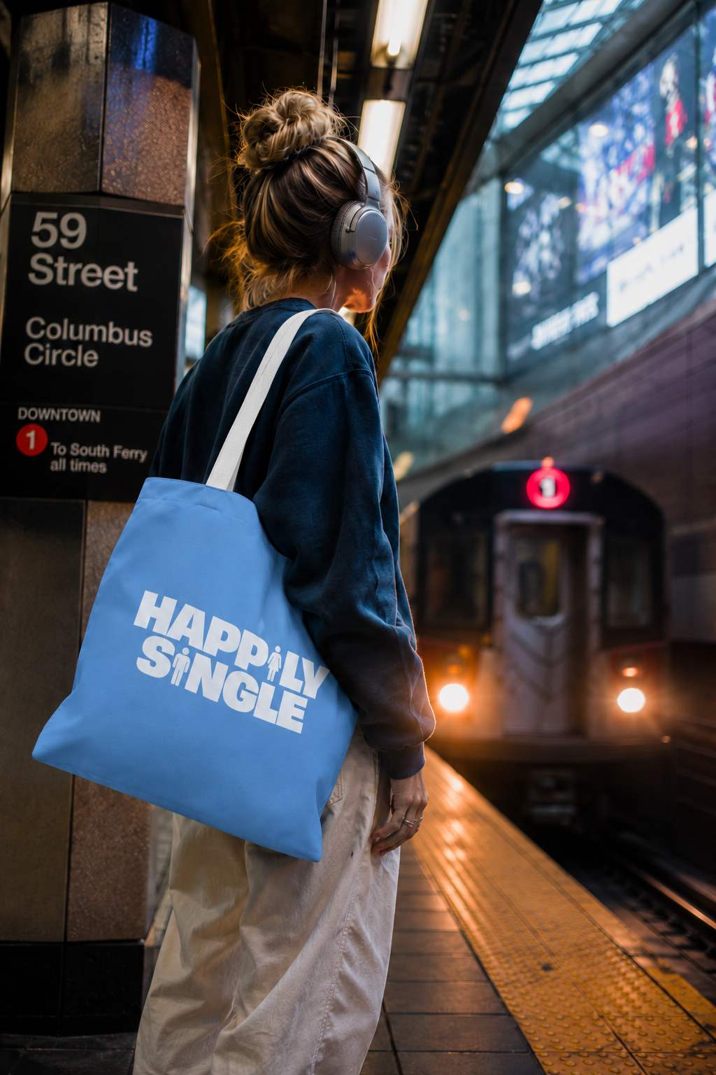 Gen Z woman carrying a blue Happily Single tote bag while waiting for the train at Columbus Circle in New York City.