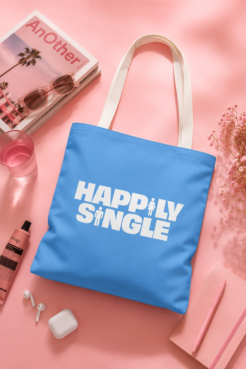 Blue Happily Single tote bag with white straps styled on a soft pastel pink background with lifestyle accessories.
