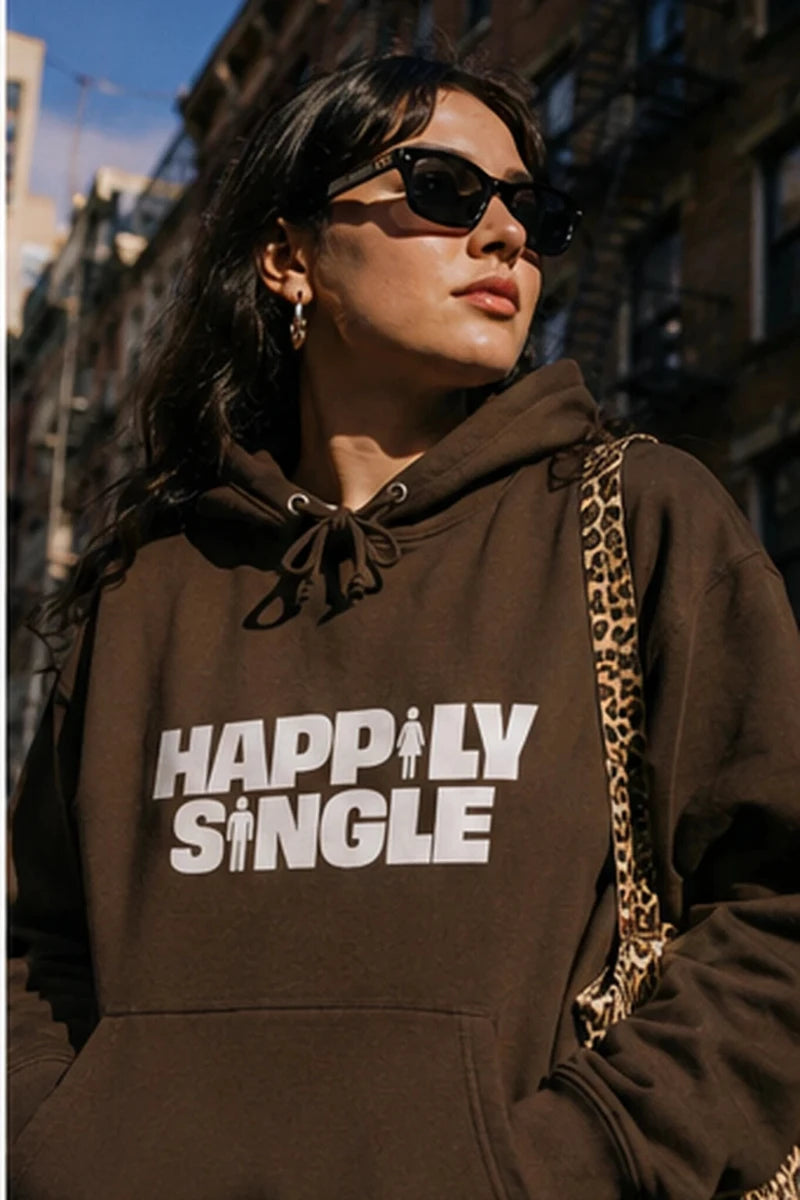 Gen Z woman wearing brown Happily Single hoodie in colorful city street setting