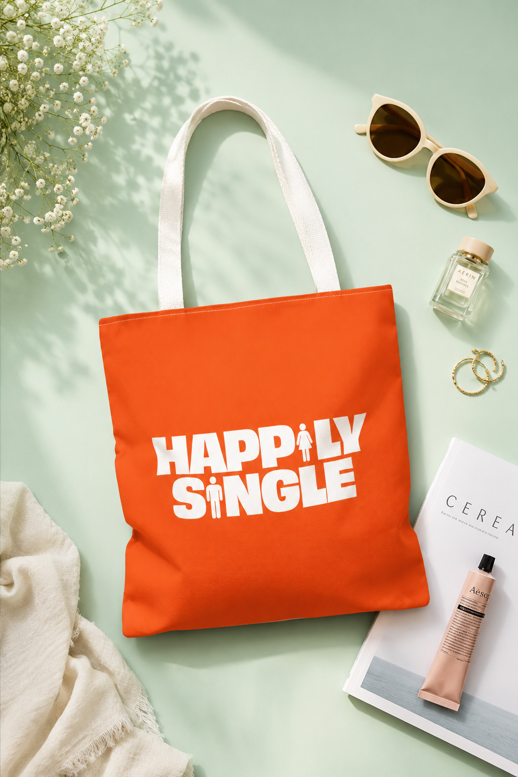 Orange Happily Single tote bag styled in an editorial flat lay on a pastel mint green background with lifestyle accessories.