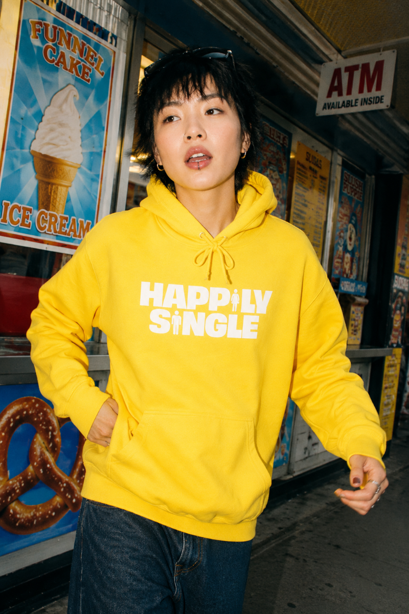 Gen Z model wearing yellow Happily Single hoodie outside colorful neighborhood shop
