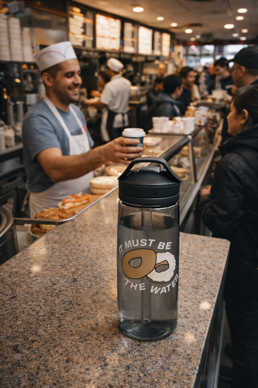 New York bagel humor water bottle 25oz reusable bottle with It Must Be The Water donut design on café counter