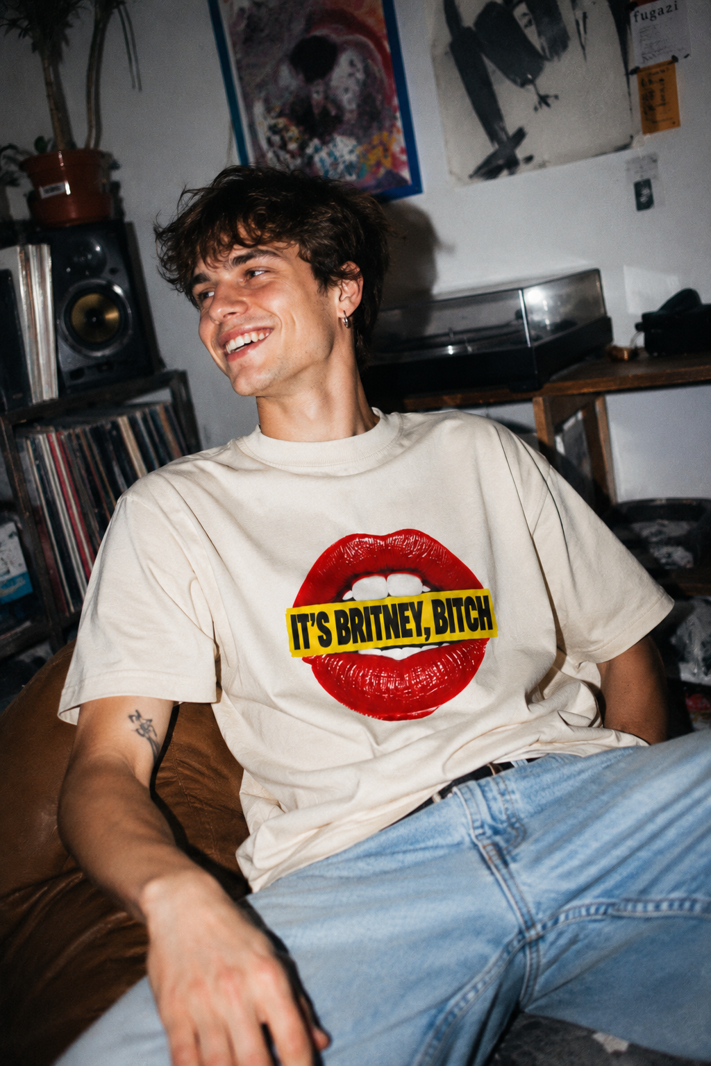 Male Gen Z wearing the cream It’s Britney, Bitch Britney Spears homage T-shirt in a candid record room scene.