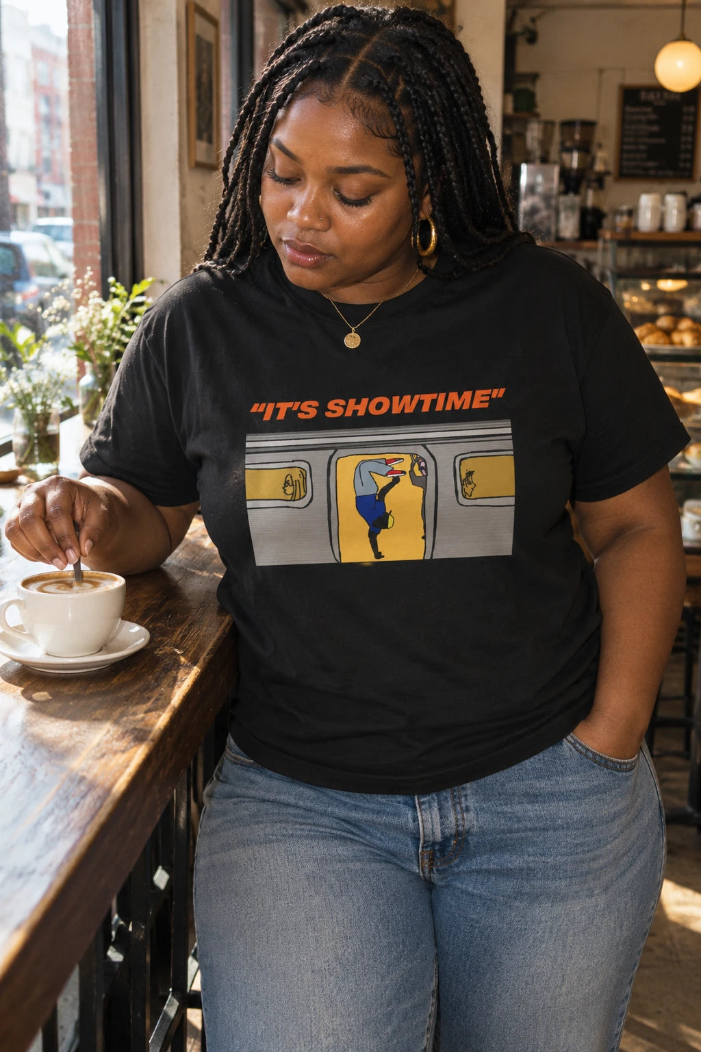 Woman wearing a black It’s Showtime NYC subway graphic T-shirt while stirring coffee at a café counter.
