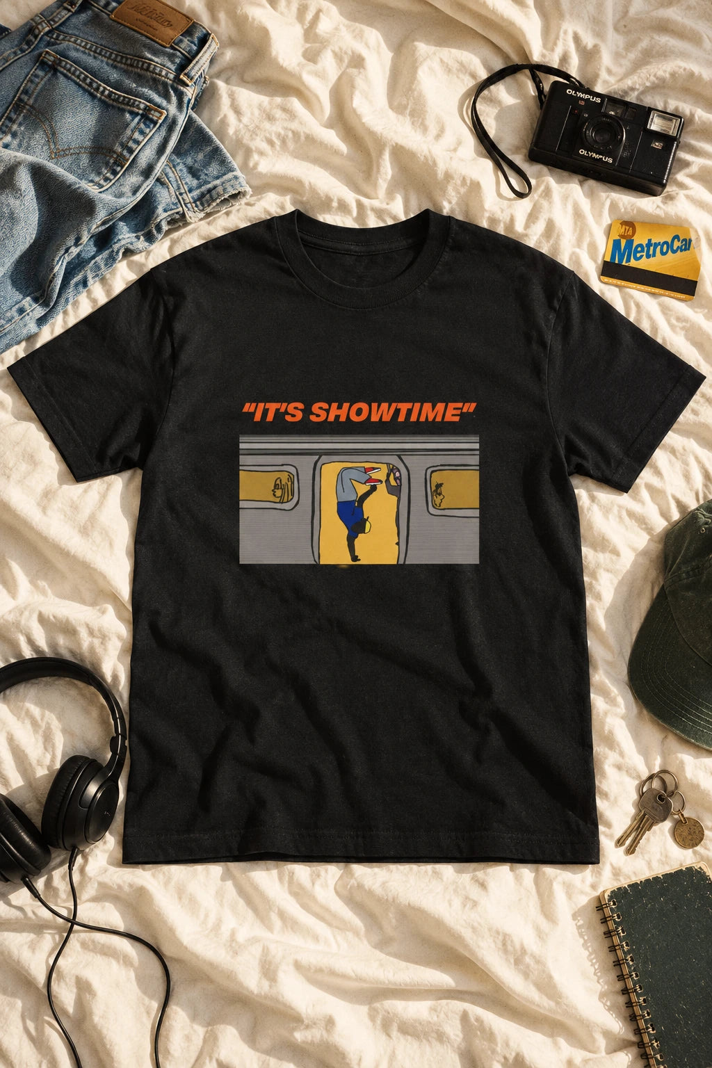 Black It’s Showtime subway graphic T-shirt laid flat on ivory bedding beside jeans, headphones and a camera.