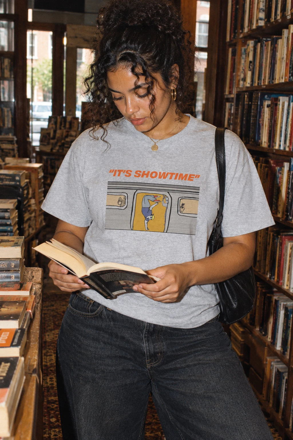 Woman reading in a bookshop while wearing a heather grey It’s Showtime NYC subway graphic T-shirt.