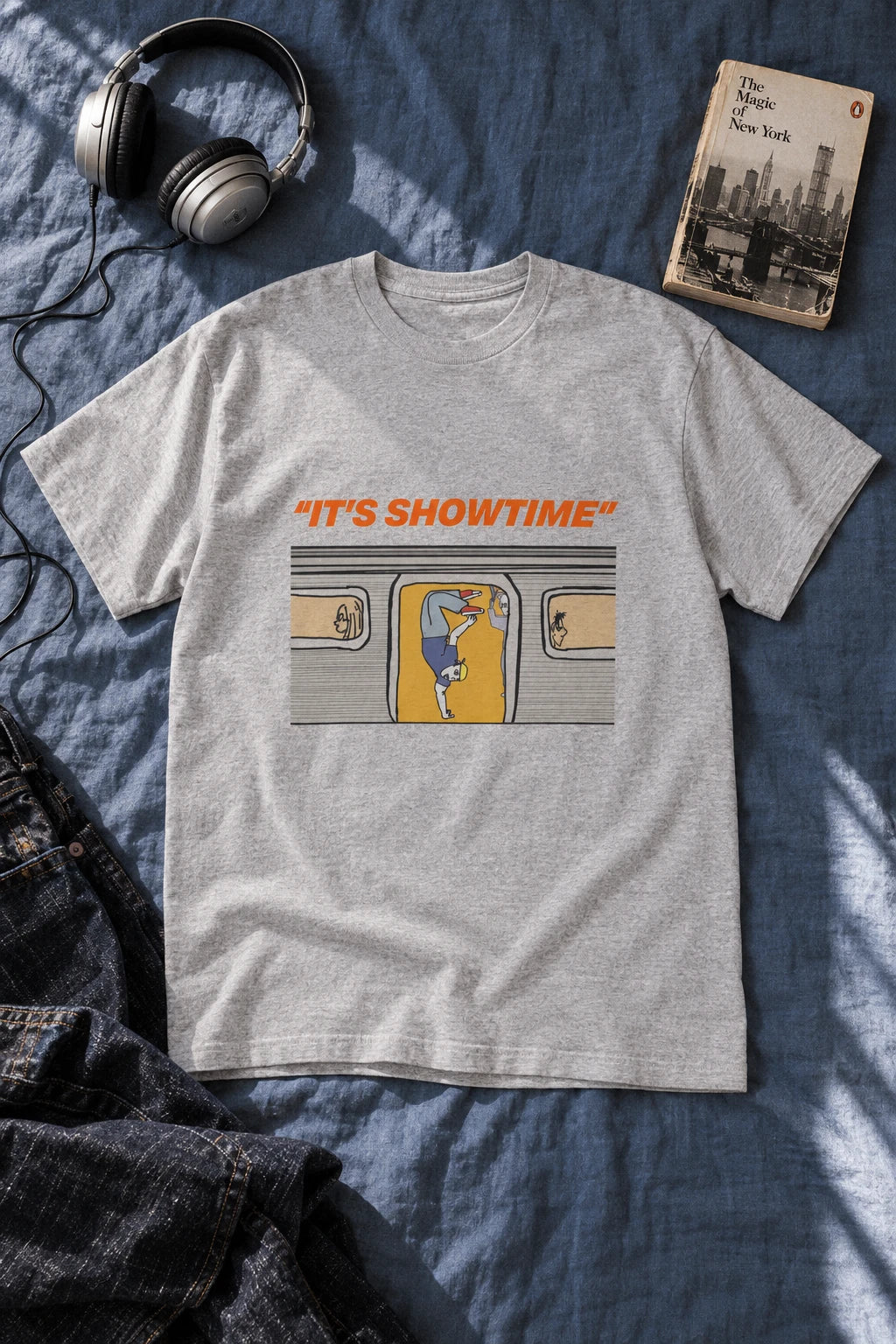 Heather grey It’s Showtime subway graphic T-shirt laid flat on blue bedding beside headphones and a book.