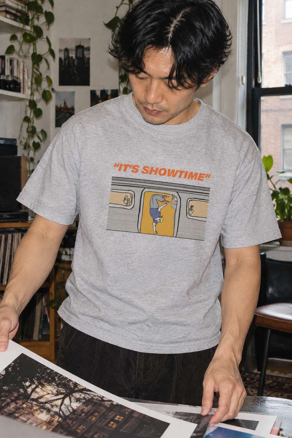 Man wearing a heather grey It’s Showtime subway graphic T-shirt while sorting photographs at a worktable.