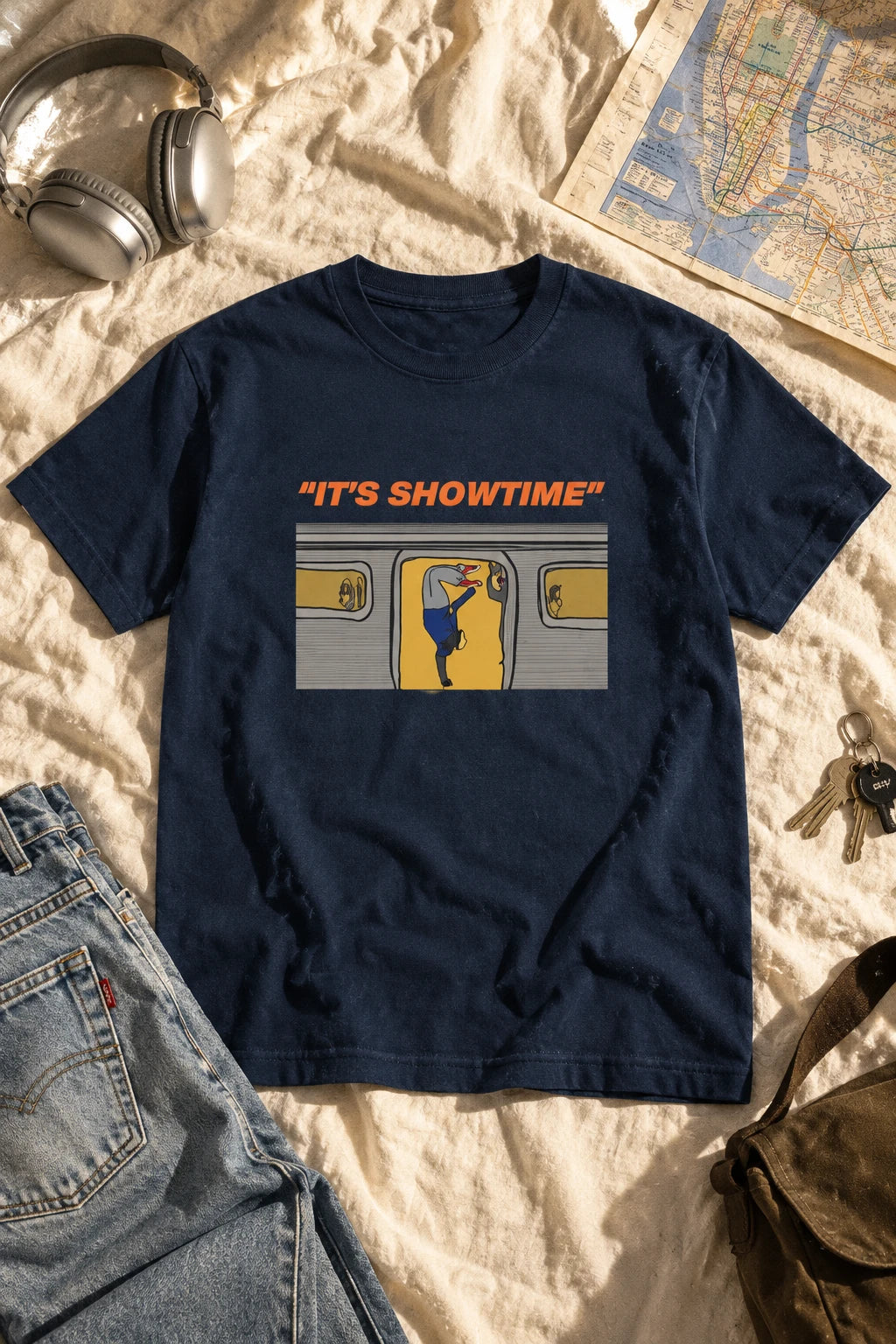 Navy It’s Showtime subway graphic T-shirt laid flat on ivory bedding beside headphones, jeans and a map.