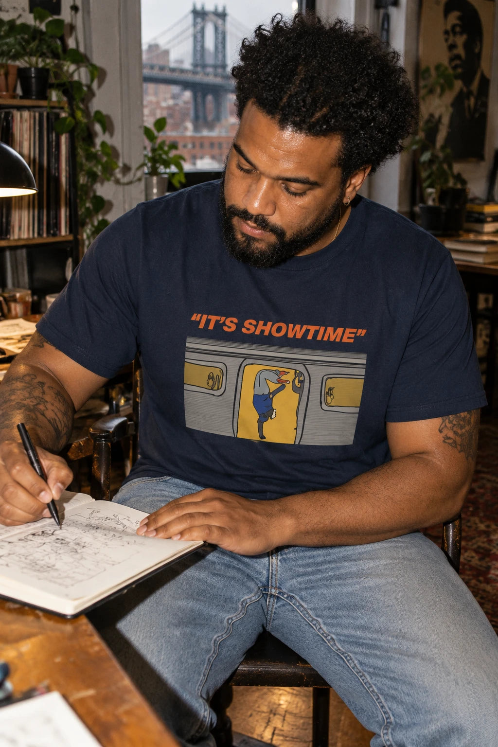 Man wearing a navy It’s Showtime subway graphic T-shirt while drawing in a sketchbook at a wooden desk.