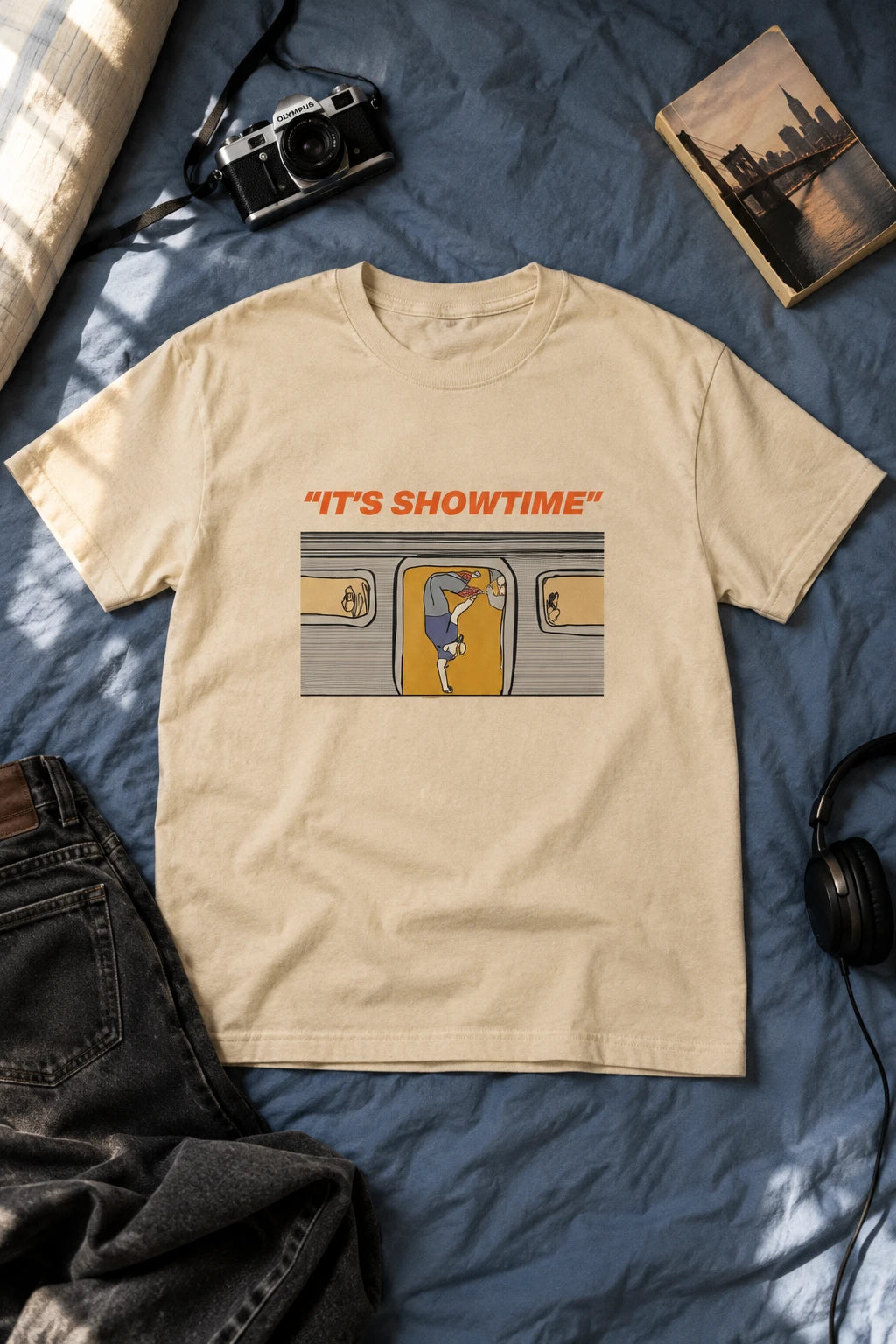 Sand It’s Showtime subway graphic T-shirt laid flat on blue bedding beside a camera, book and jeans.