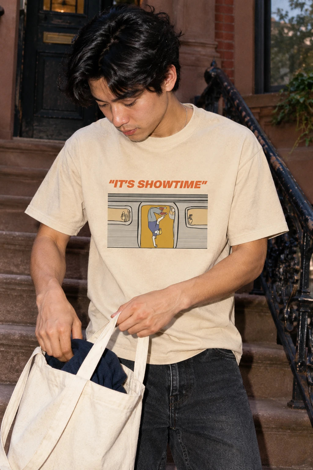 Man wearing a sand It’s Showtime NYC subway T-shirt while checking a canvas tote on a brownstone stoop.