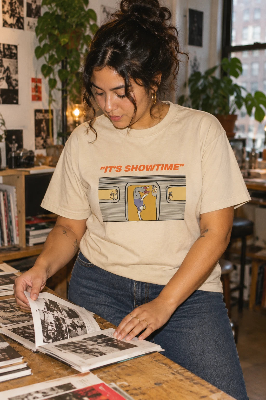 Woman wearing a sand It’s Showtime subway graphic T-shirt while browsing a photo book at a studio table.