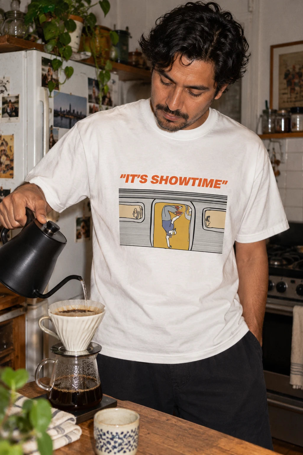 Man wearing a white It’s Showtime NYC subway graphic T-shirt while making pour-over coffee.