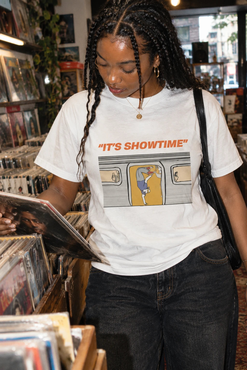 Woman browsing records while wearing a white It’s Showtime T-shirt with a subway dancer illustration.