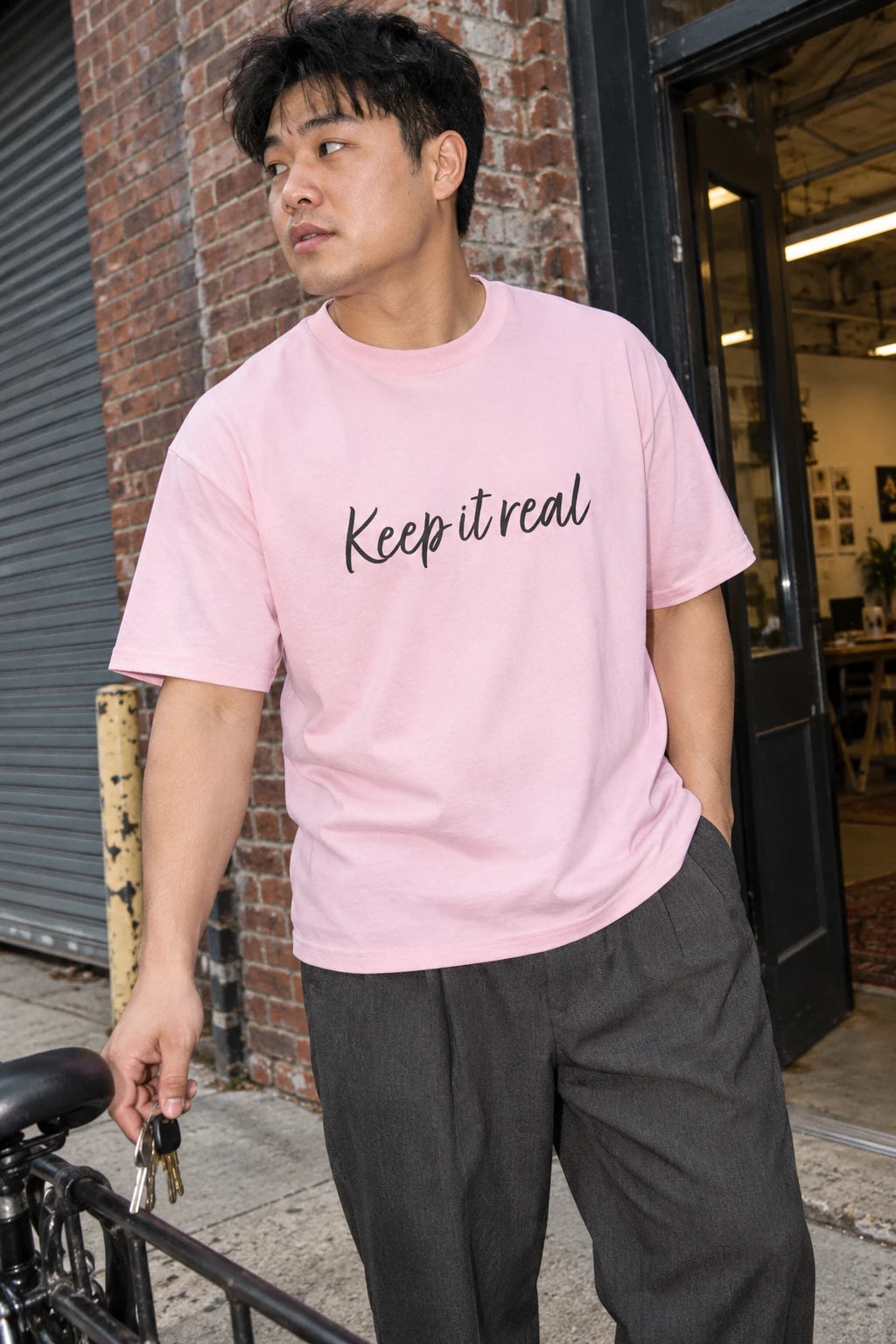 Man wearing Tantrum Media’s light pink Keep It Real T-Shirt beside a bicycle outside a brick building.