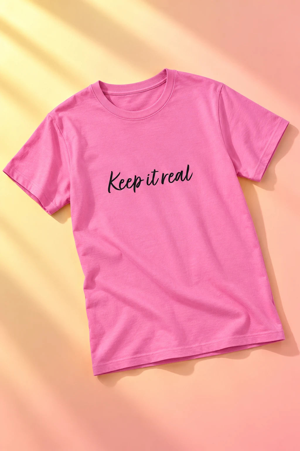 Tantrum Media pink Keep It Real T-Shirt with black script lettering, laid flat on a peach and yellow gradient background.