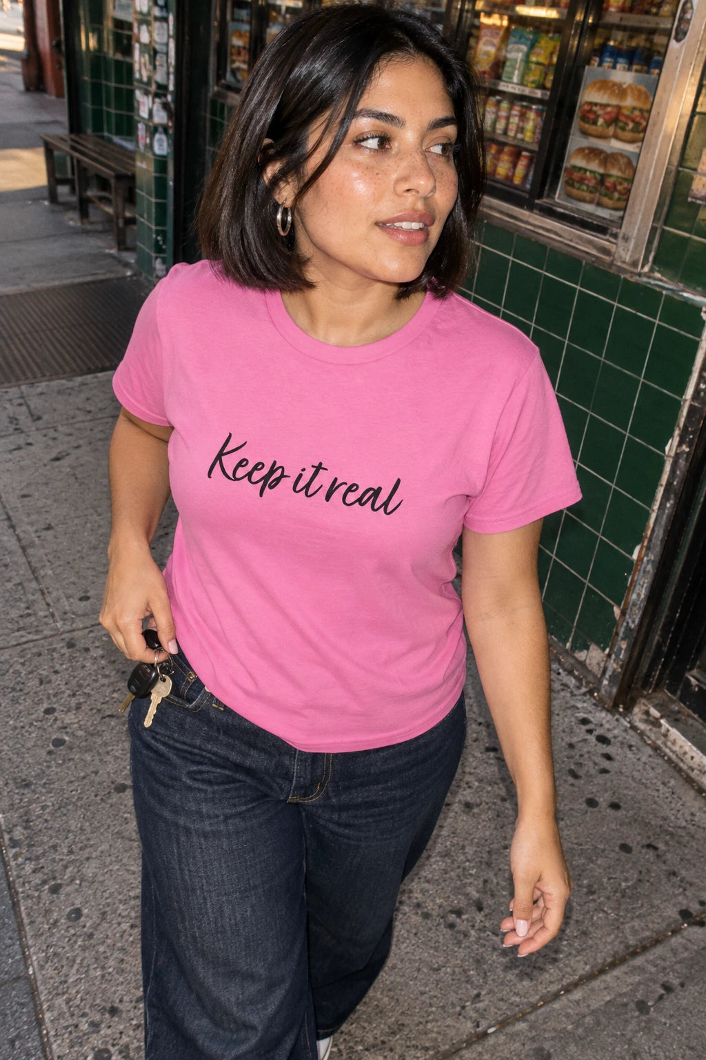 Woman wearing Tantrum Media’s pink Keep It Real T-Shirt with black script lettering outside a city deli.