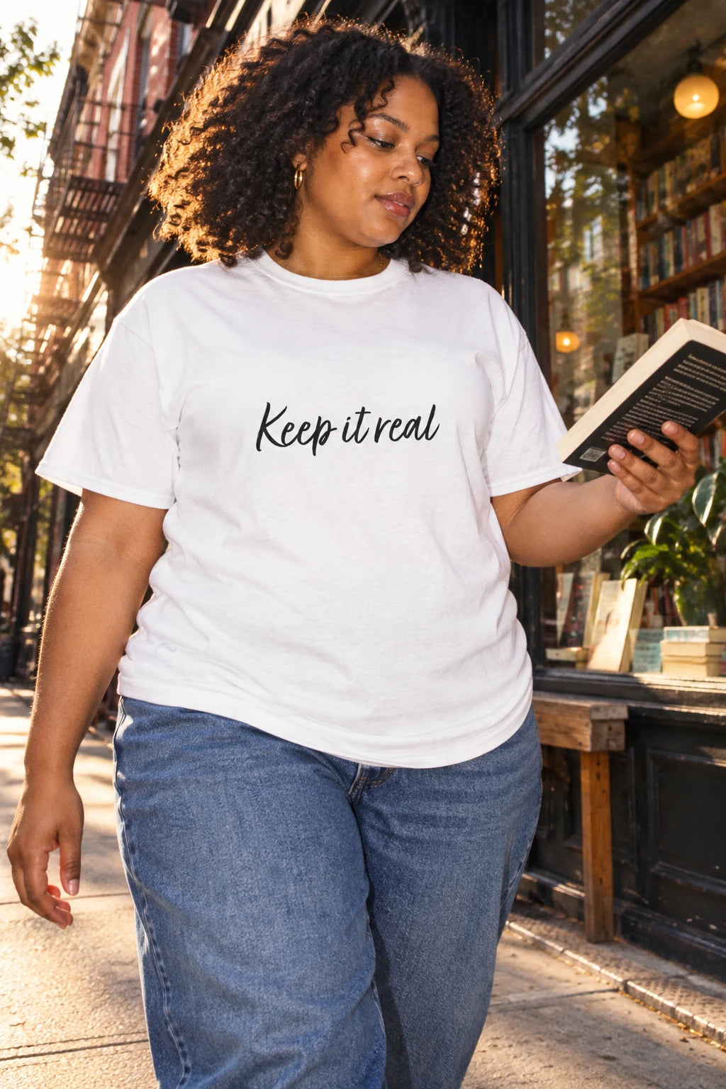 Woman reading a book outside a bookstore in Tantrum Media’s white Keep It Real T-Shirt with black script lettering.