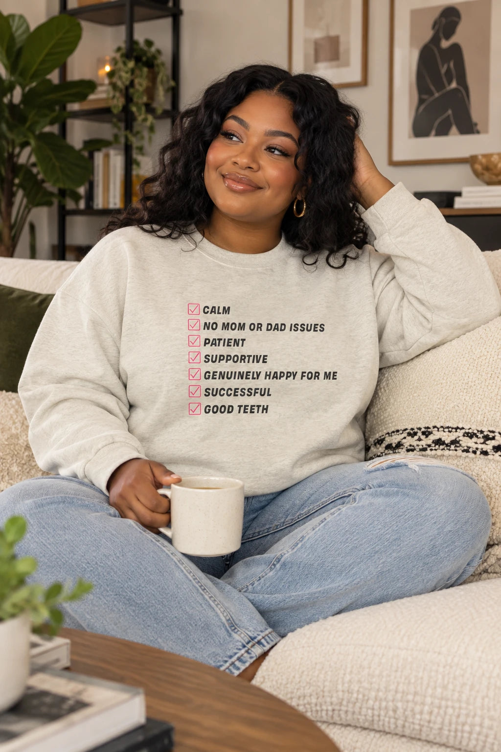 Oatmeal Kim Kardashian’s Men’s Must-Have List crewneck sweatshirt on a curvy woman sitting on a couch in a cozy living room.