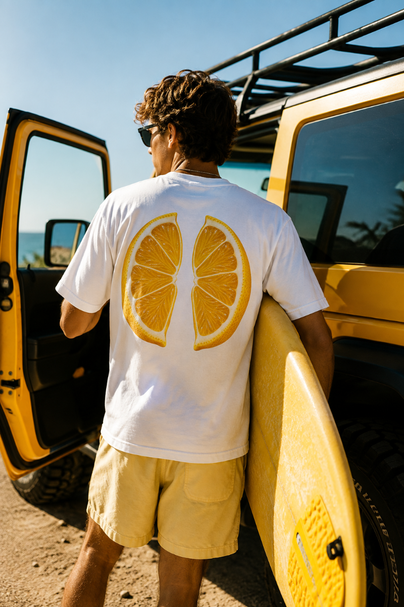 Male wearing white lemon graphic back print T-shirt with yellow surfboard beside a Jeep