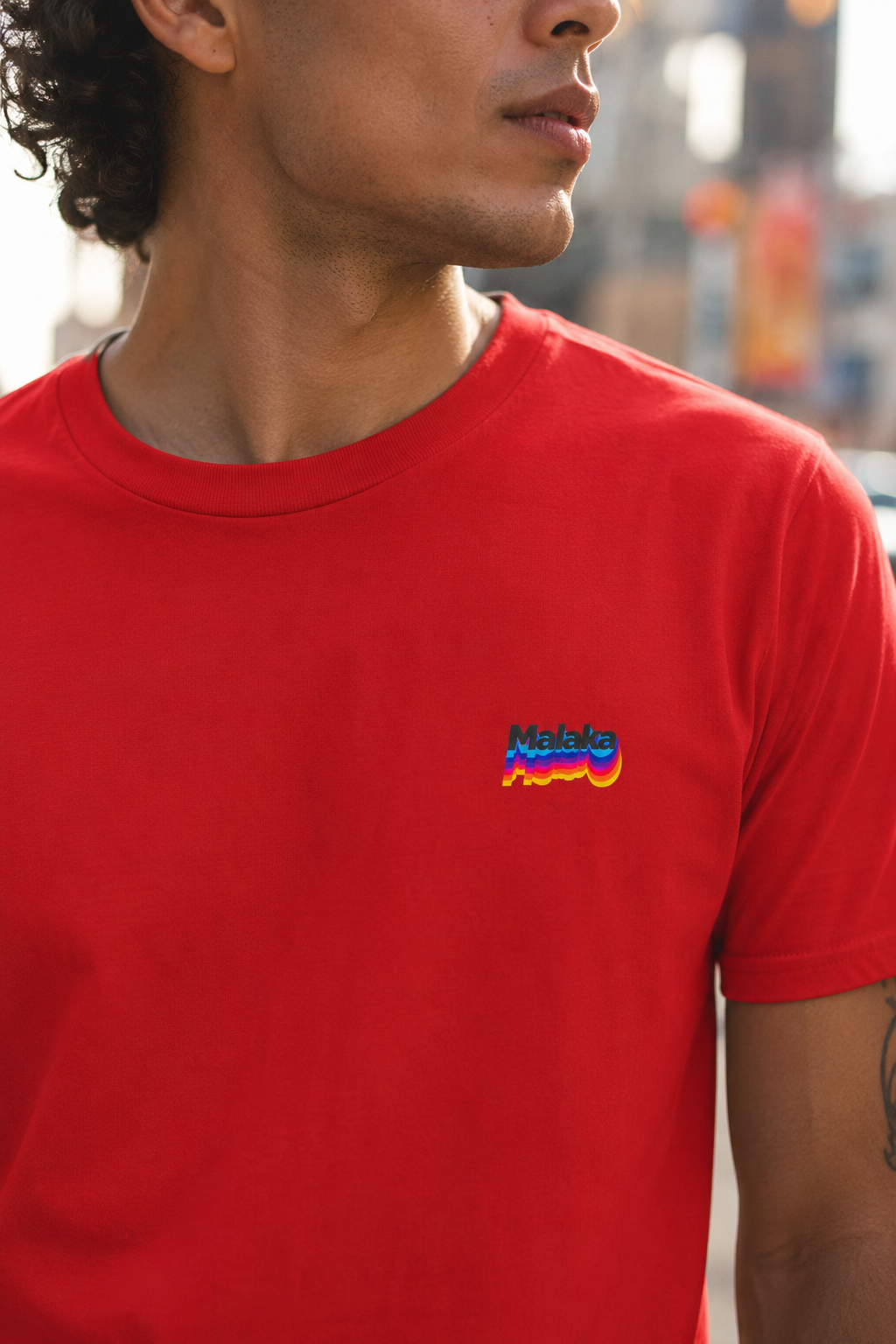 Man wearing a red Malaka Greek slang T-shirt with a small colorful chest graphic in an urban setting