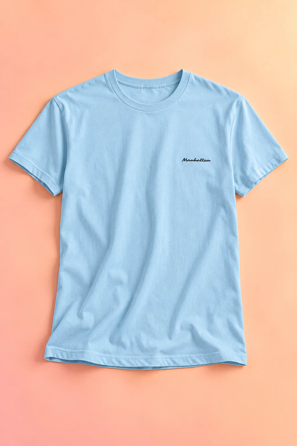 Light blue Manhattan script T-shirt laid flat on a soft peach gradient background.