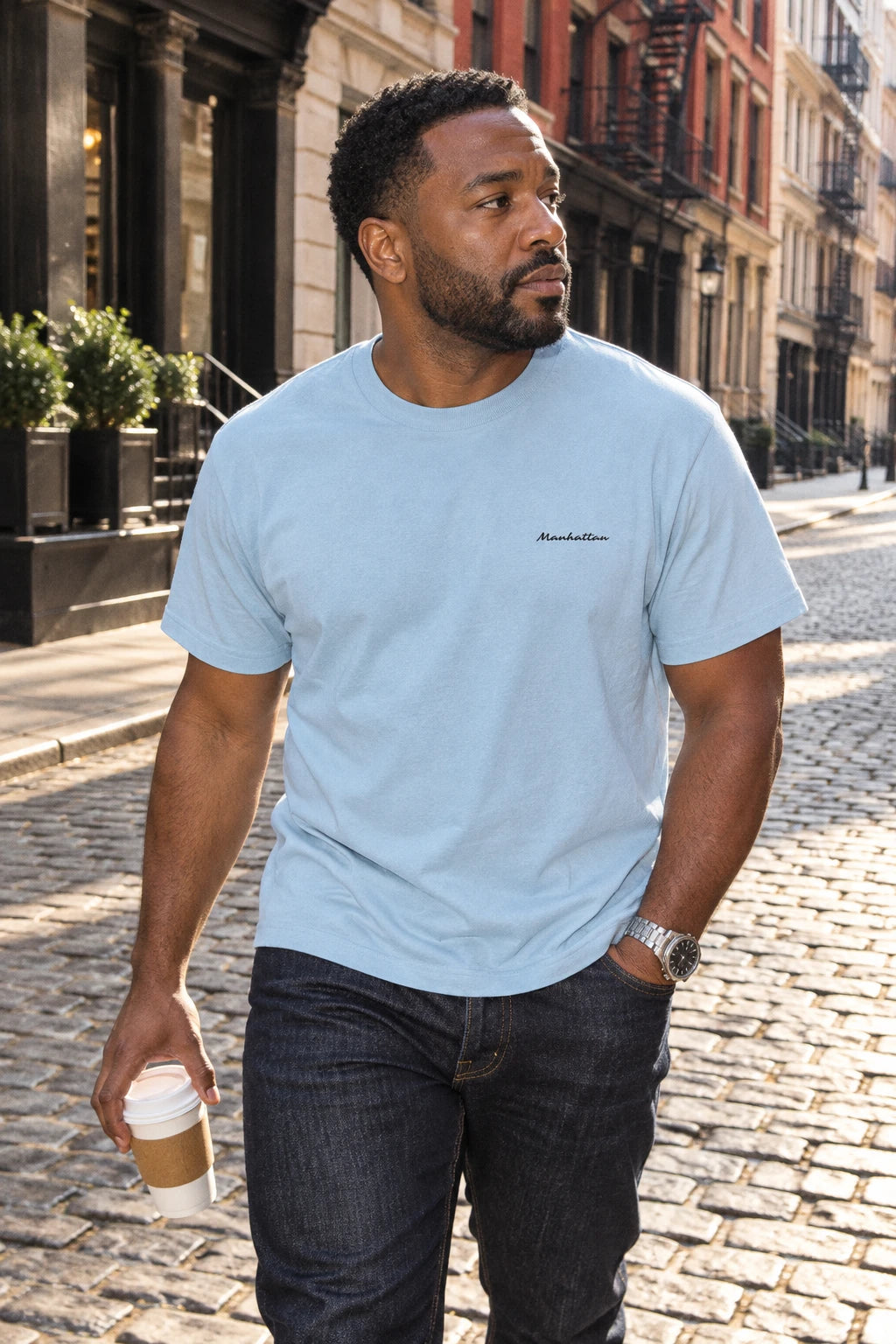 Man wearing a light blue Manhattan T-shirt while carrying coffee along a cobblestone street in Tribeca.