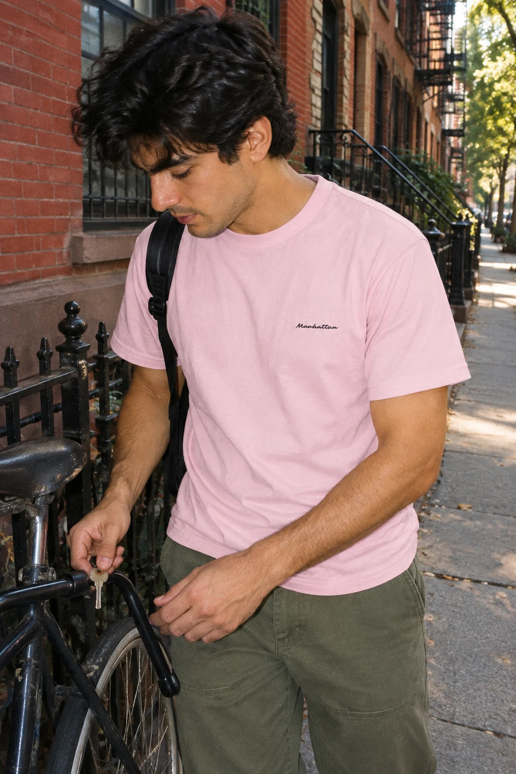 Man wearing a pink Manhattan T-shirt while unlocking a bicycle beside a brick building.
