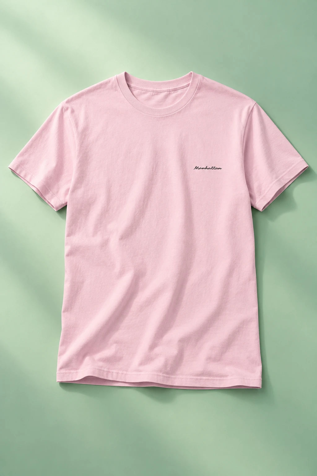 Pink Manhattan script T-shirt laid flat on a soft mint-green gradient background.