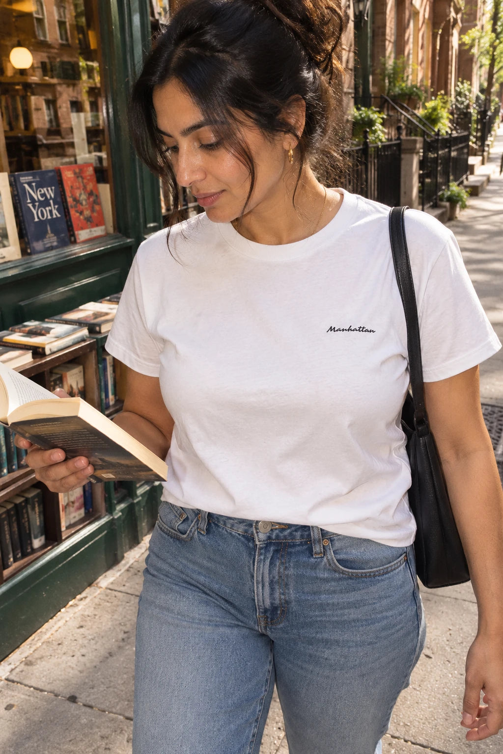 Woman wearing a white Manhattan script T-shirt and jeans while reading outside a neighborhood bookstore.