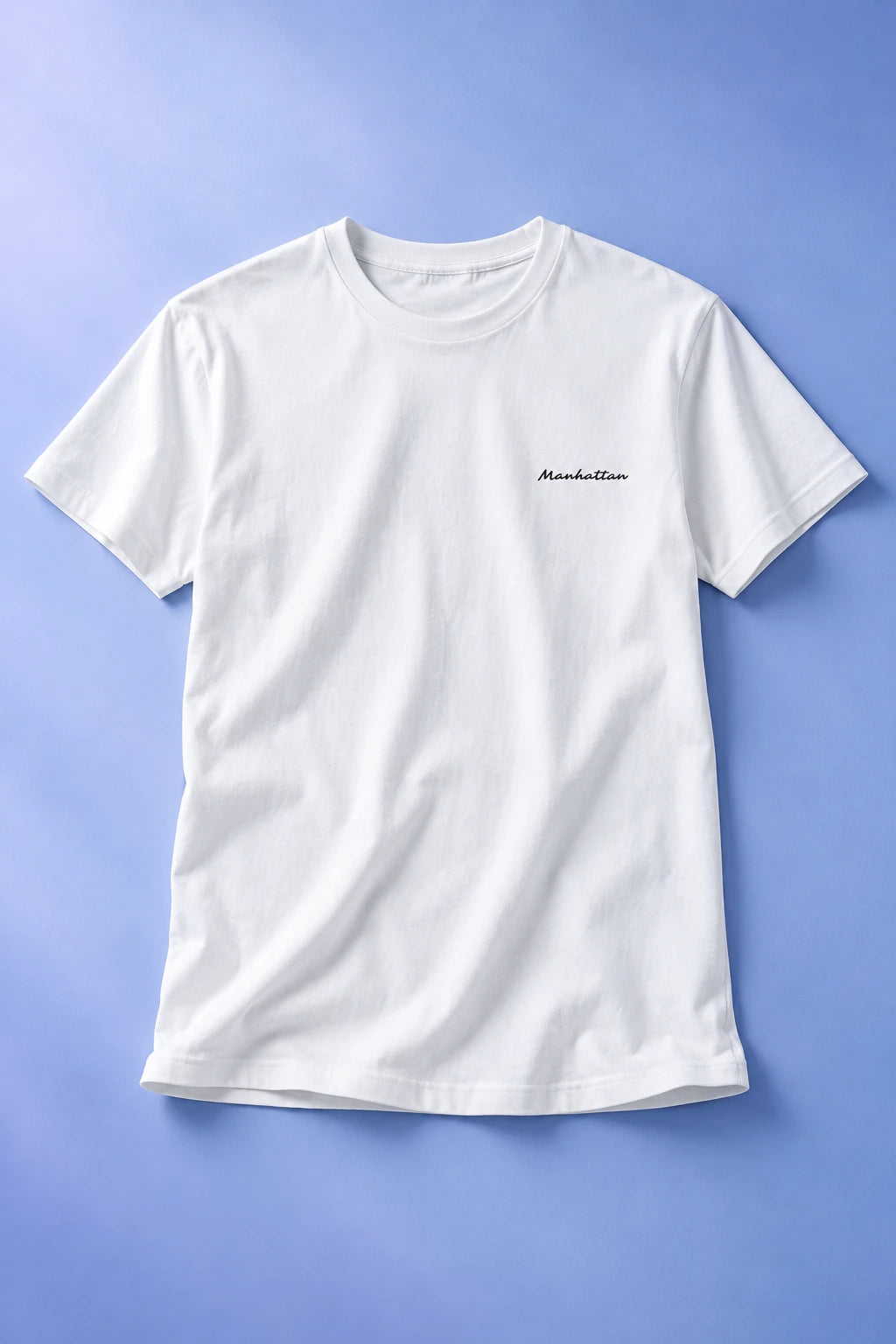 White Manhattan T-shirt laid flat on a soft periwinkle gradient background, showing the small black script chest print.