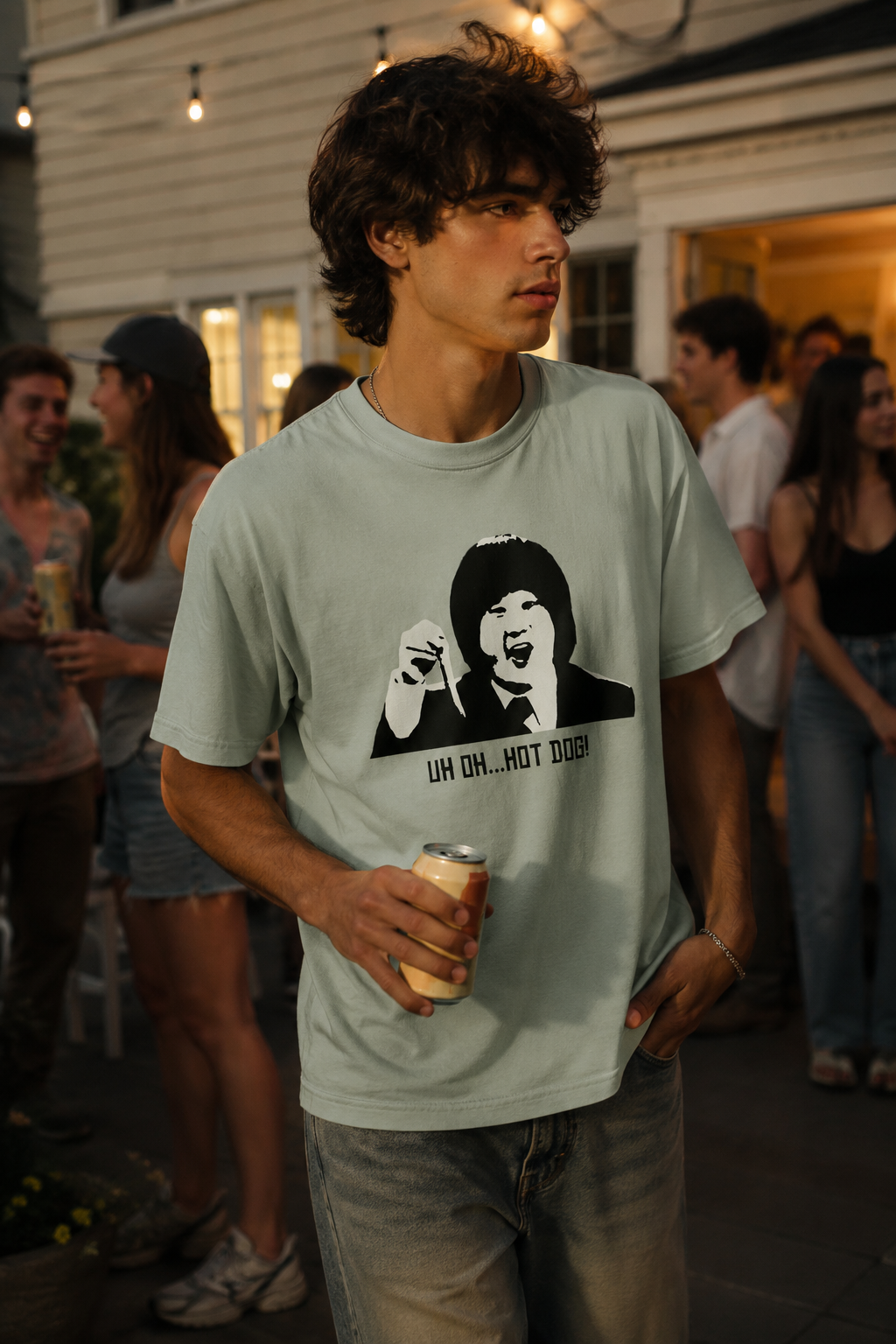 Young man wearing a sage green graphic T-shirt with a retro portrait print and "UH OH... HOT DOG!" text while holding a drink at an outdoor gathering.