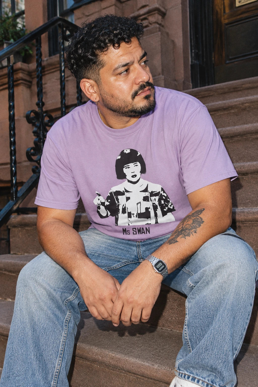 Man wearing a lavender Tantrum Media Ms Swan graphic T-shirt and light blue jeans, seated on brownstone steps.