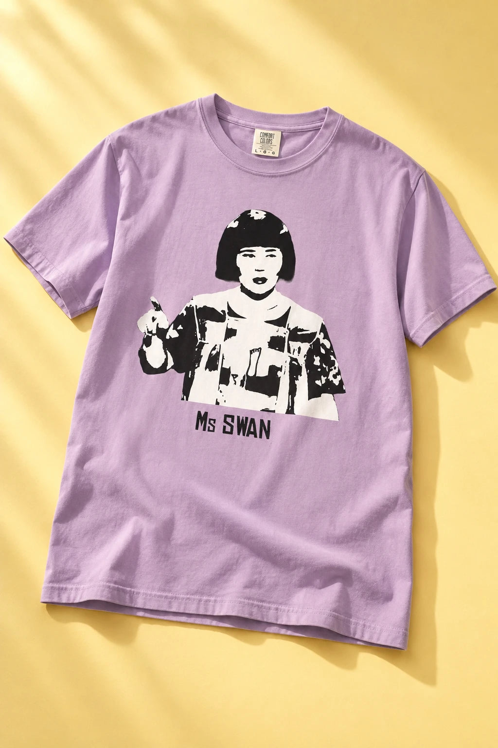 Tantrum Media lavender Ms Swan T-Shirt laid flat on a pale yellow background with soft folds and directional sunlight.