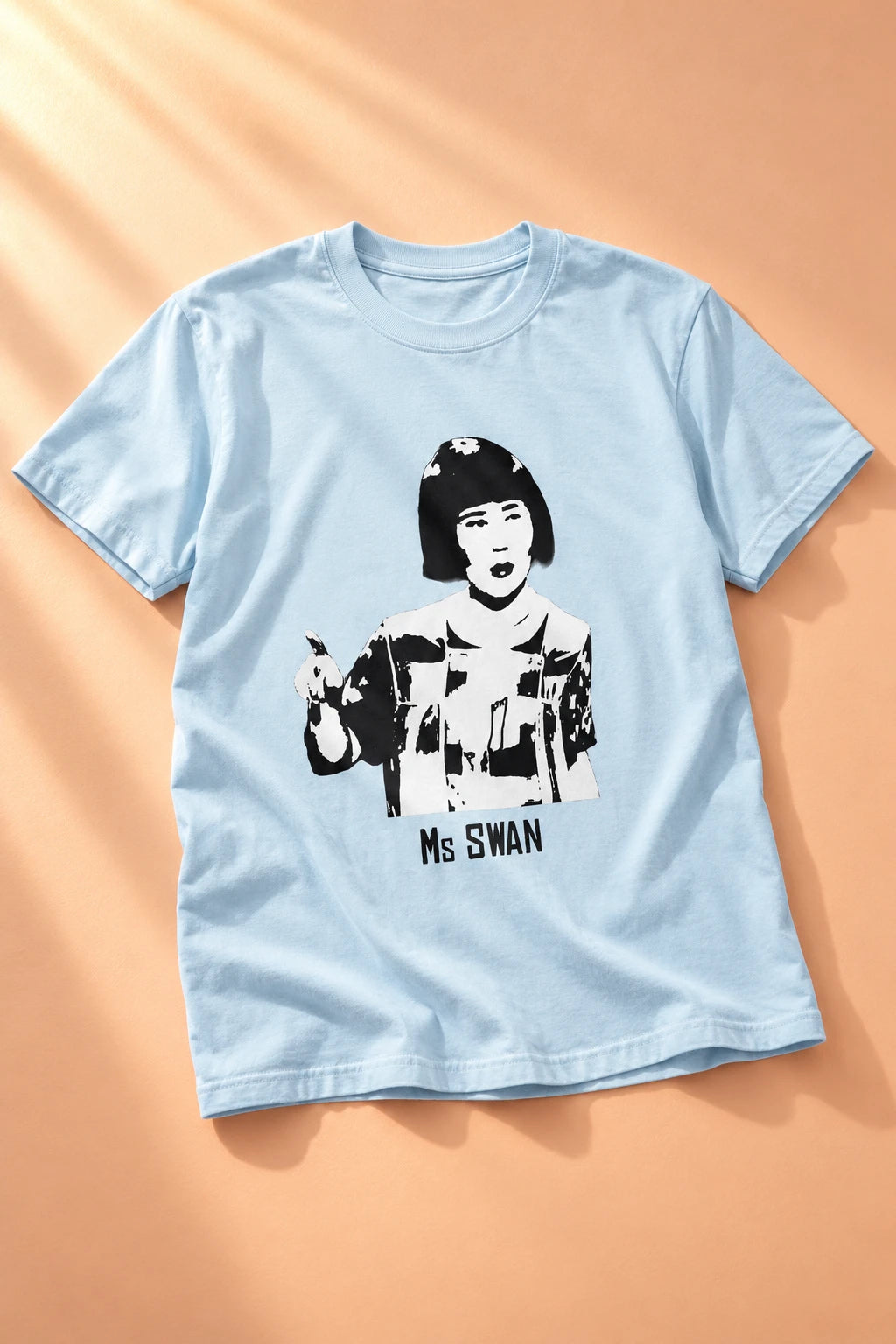Tantrum Media light blue Ms Swan graphic T-shirt laid flat on a peach background in warm directional sunlight.