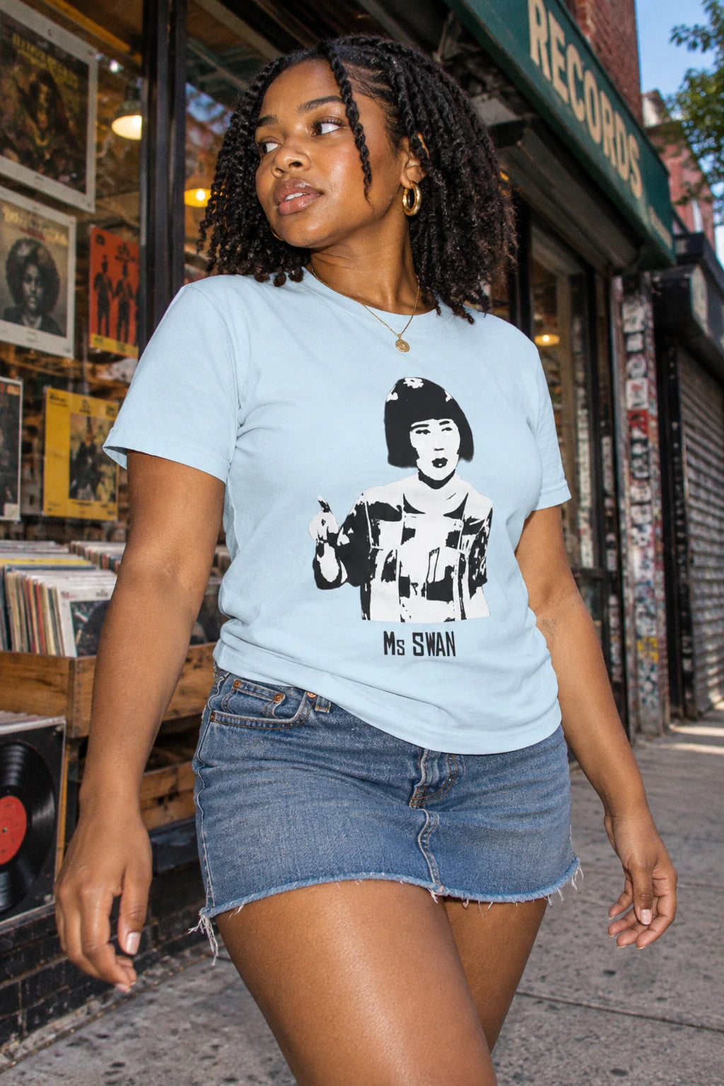 Woman wearing a light blue Tantrum Media Ms Swan T-Shirt with a denim miniskirt outside a record store.
