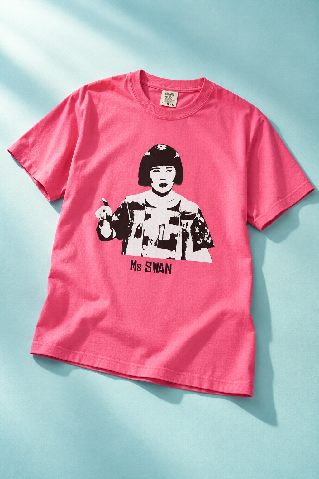 Tantrum Media bright pink Ms Swan graphic T-shirt laid flat on an aqua background with diagonal sunlight.