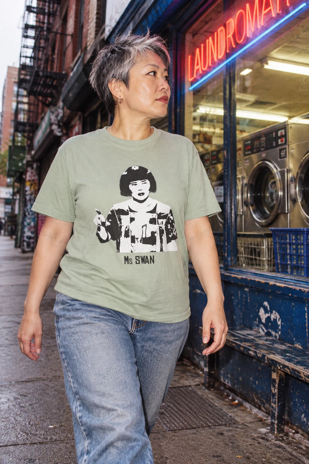 Woman with short gray hair wearing a sage green Tantrum Media Ms Swan T-Shirt and jeans outside a city laundromat.