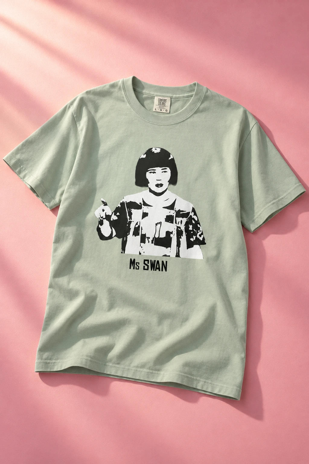 Tantrum Media sage green Ms Swan graphic T-shirt laid flat on a pink background with diagonal light and soft shadows.