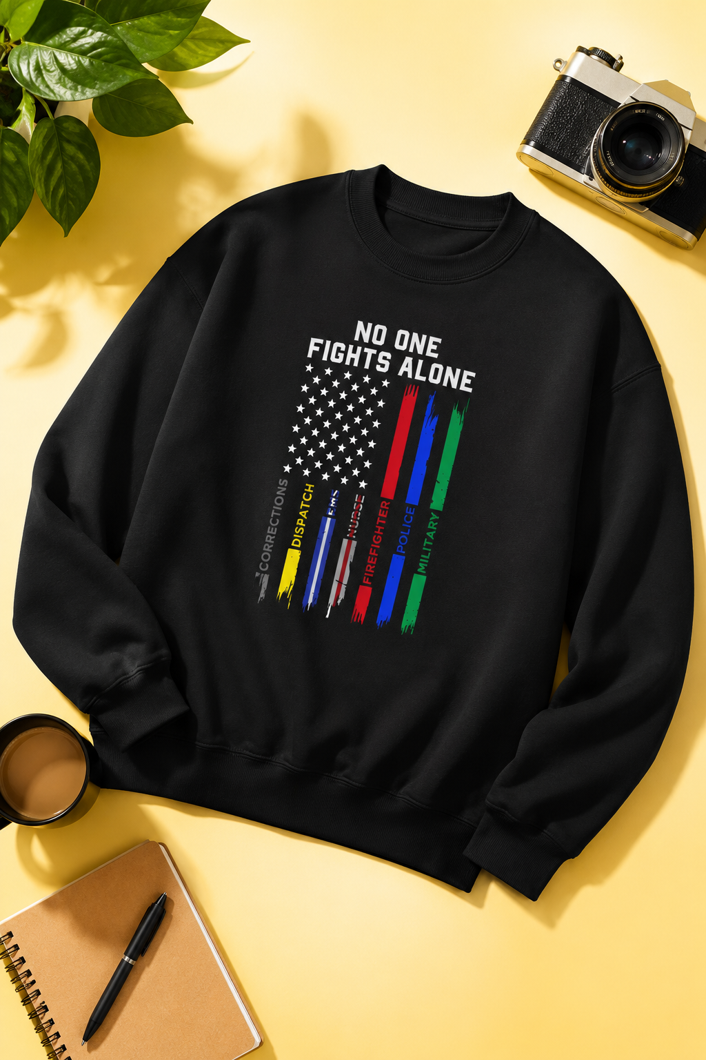 Black No One Fights Alone Workers United sweatshirt flat-lay on a soft yellow background with notebook, camera, coffee cup, and plant accents, featuring a distressed American flag design honoring corrections officers, dispatchers, EMS, nurses, firefighters, police, and military personnel.