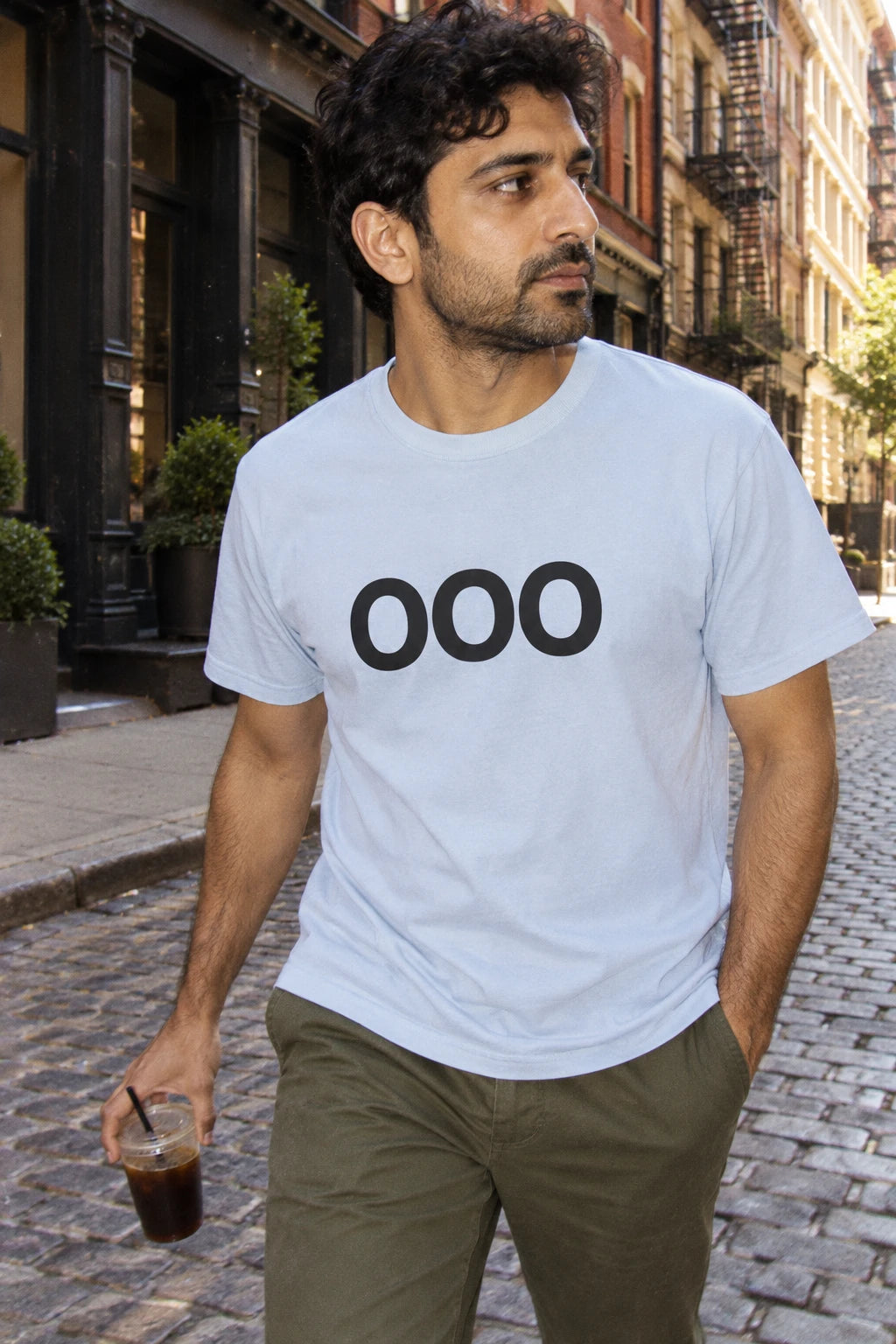 Man wearing a light blue OOO T-shirt and olive trousers while carrying iced coffee along a cobblestone street.