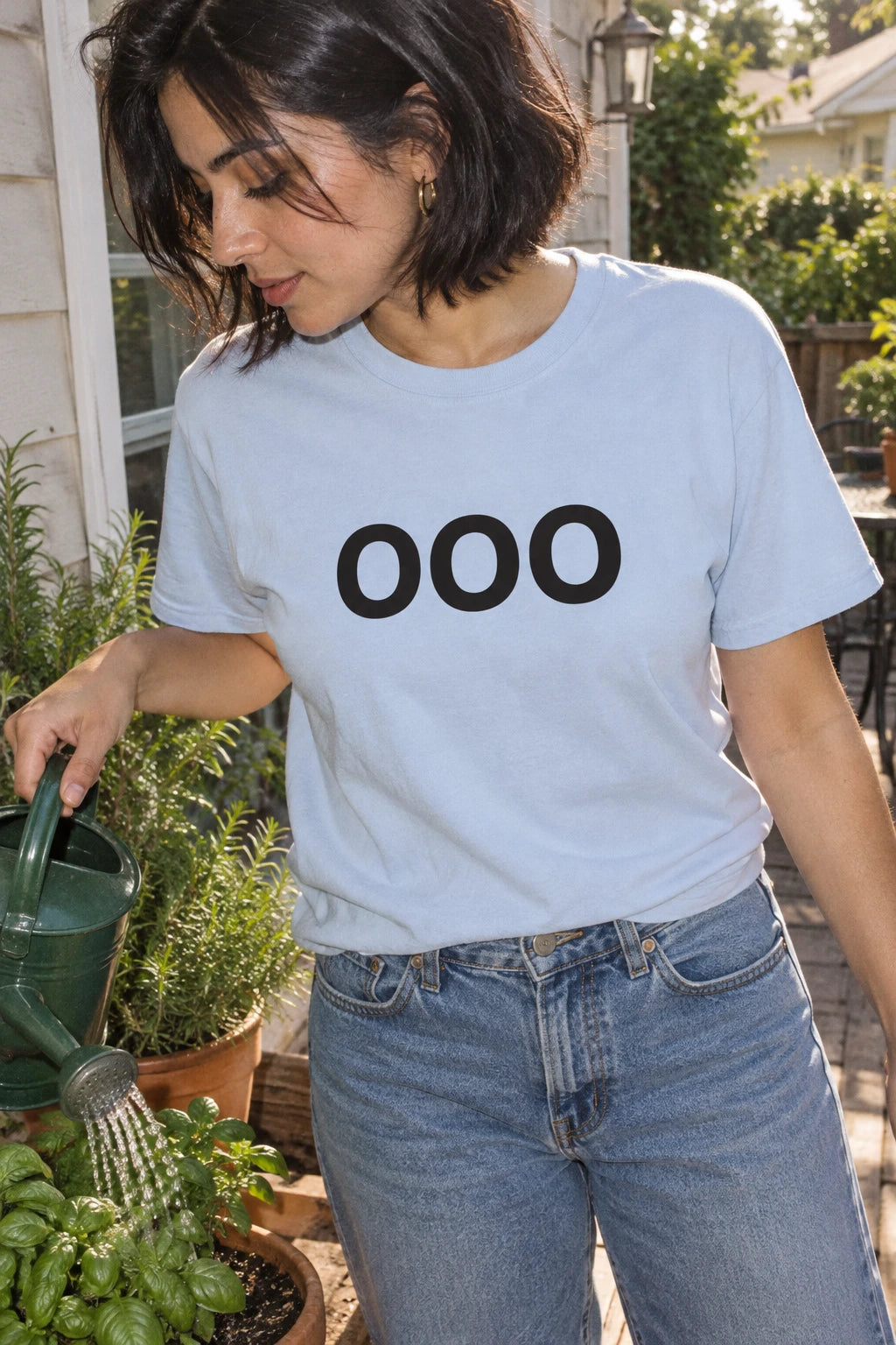 Woman wearing a light blue OOO T-shirt while watering potted herbs on a suburban porch.