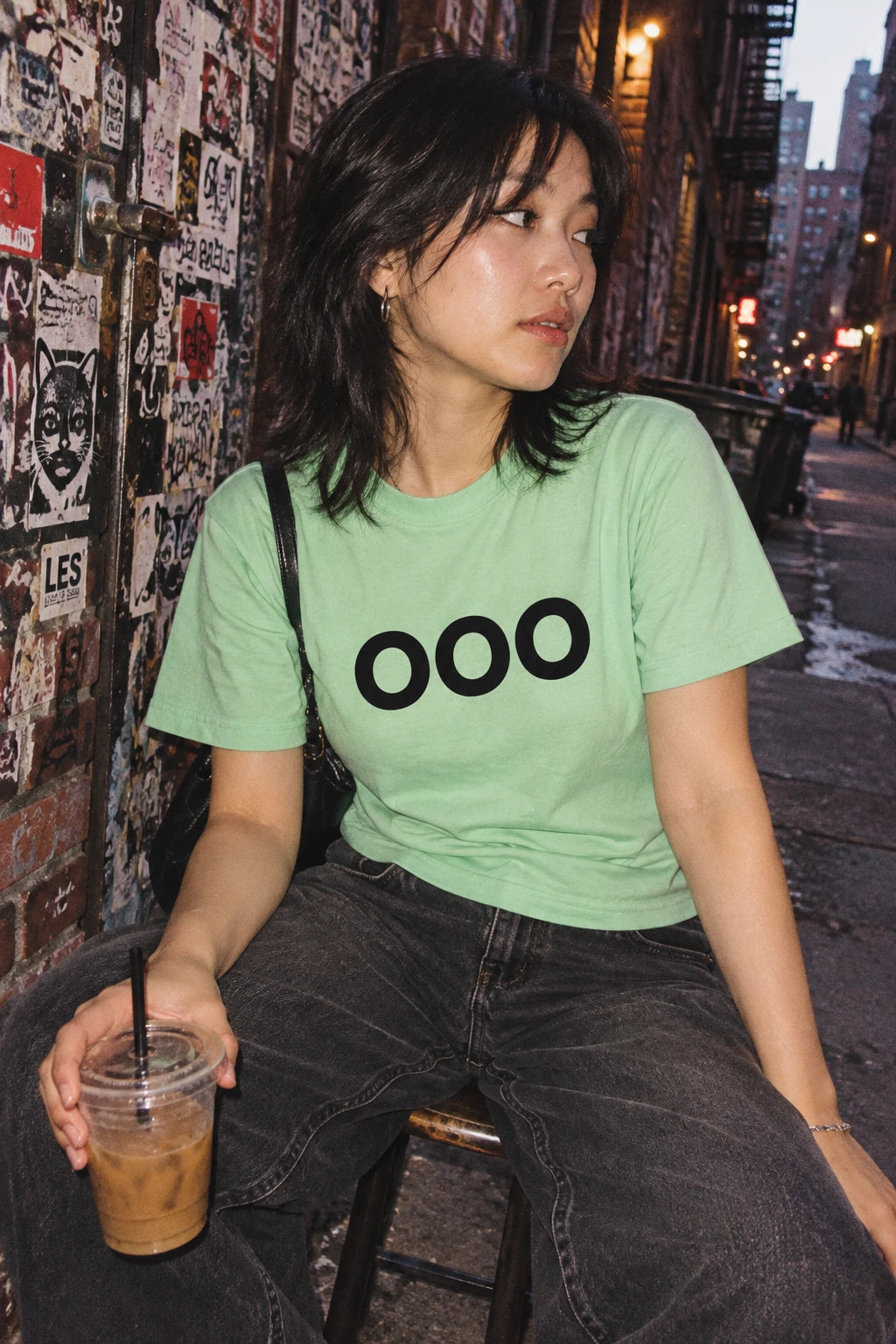 Woman wearing a mint green OOO T-shirt while sitting with an iced drink in a sticker-covered alley.
