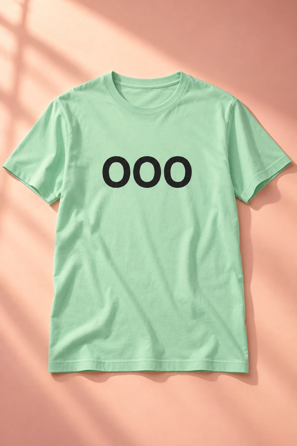Mint green OOO T-shirt laid flat on a peach background with soft window shadows.