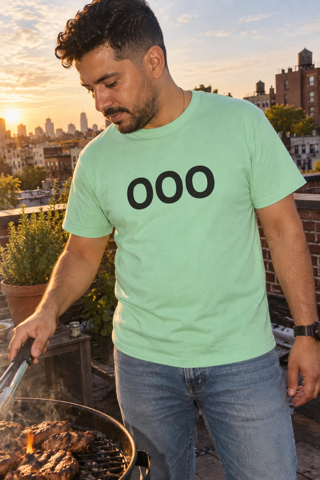 Man wearing a mint green OOO T-shirt while tending a barbecue on a Brooklyn rooftop at sunset.