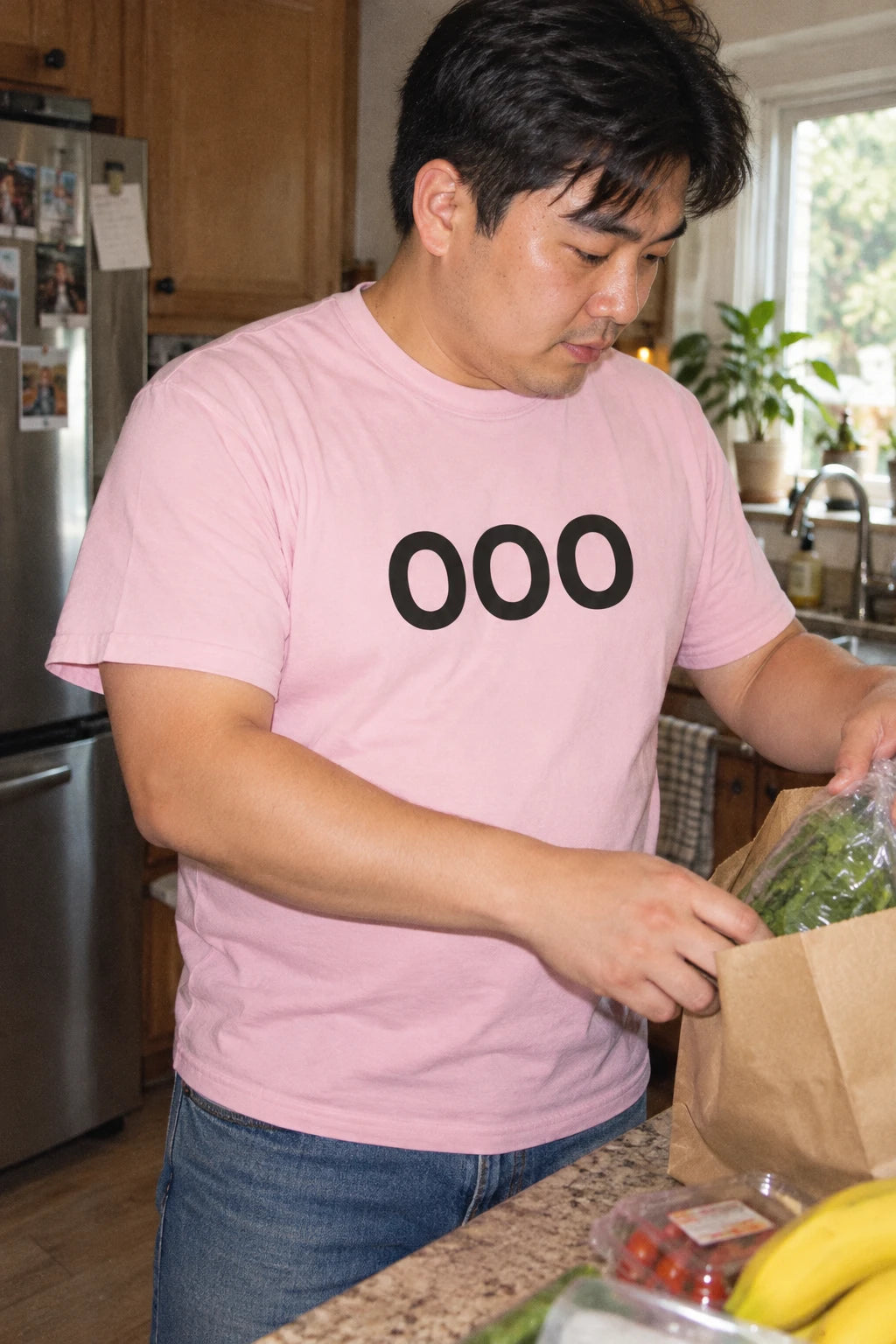 Man wearing a pink OOO T-shirt while unpacking groceries at a kitchen counter.