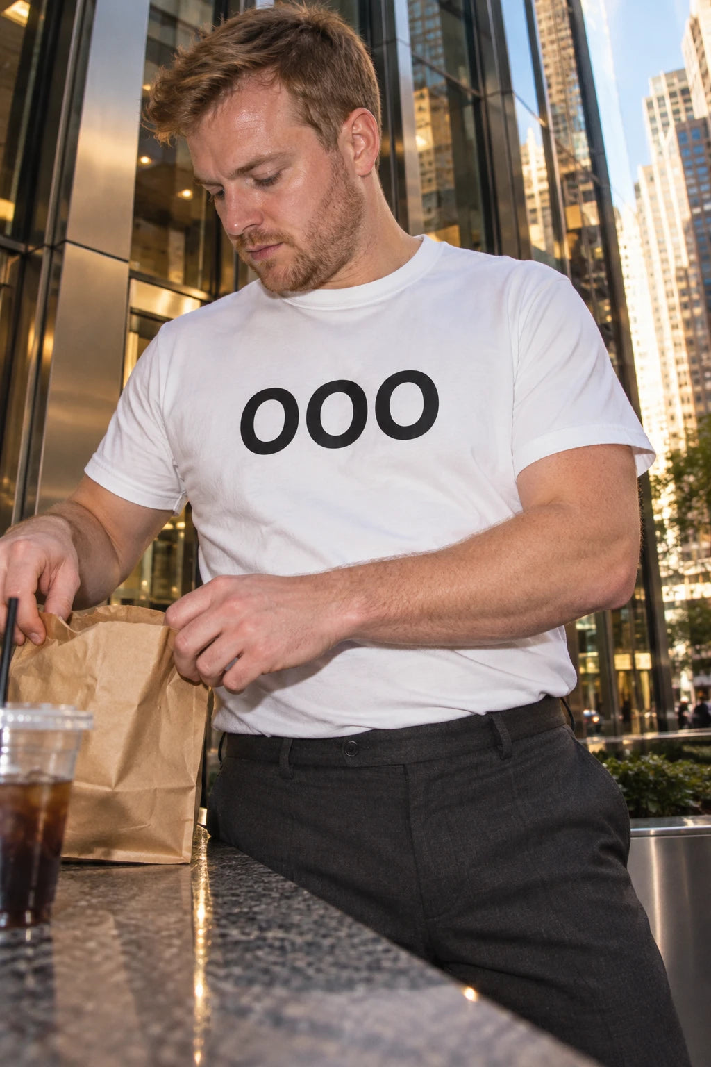 Man wearing a white OOO T-shirt and tailored trousers while opening a lunch bag outside an office building.